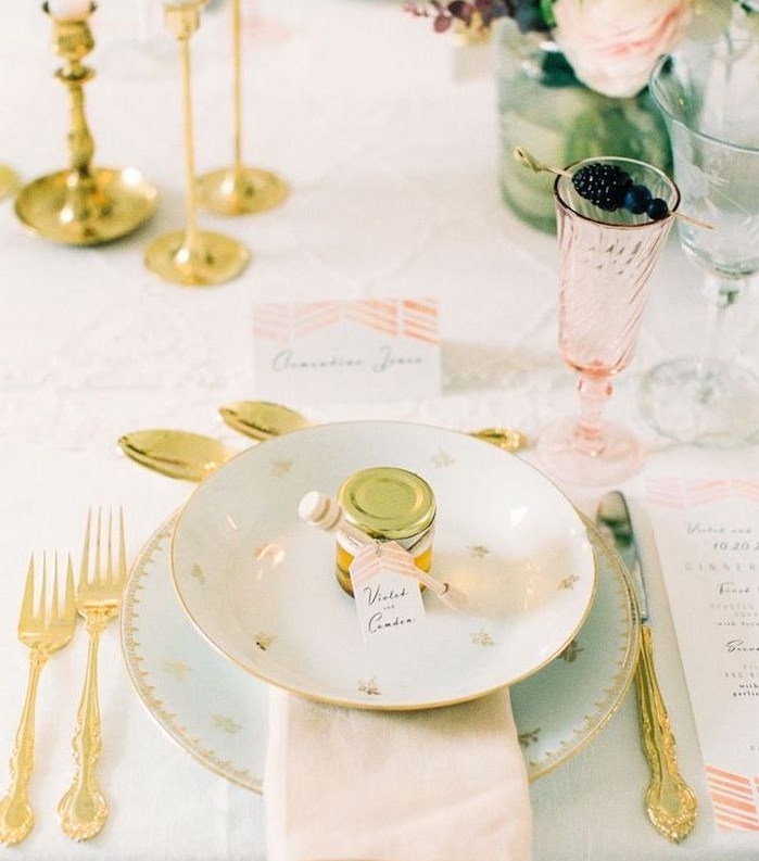 petit pot de miel dans assiettes, couverts dorés et centre de table floral, nappe blanche, verre rose transparente