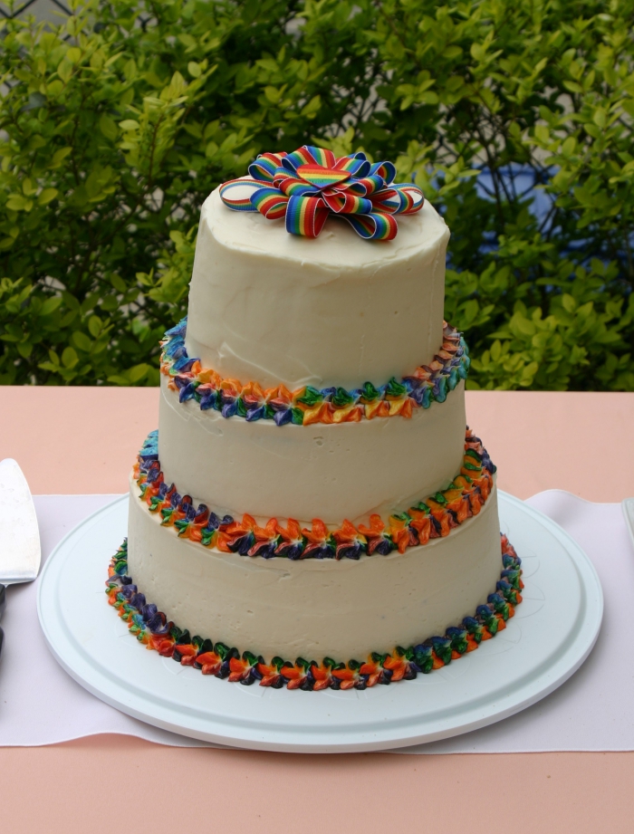 recette de wedding cake arc-en-ciel à trois étages décoré et surmonté d'un ruban multicolore décoré de glaçage multicolore sur les bords
