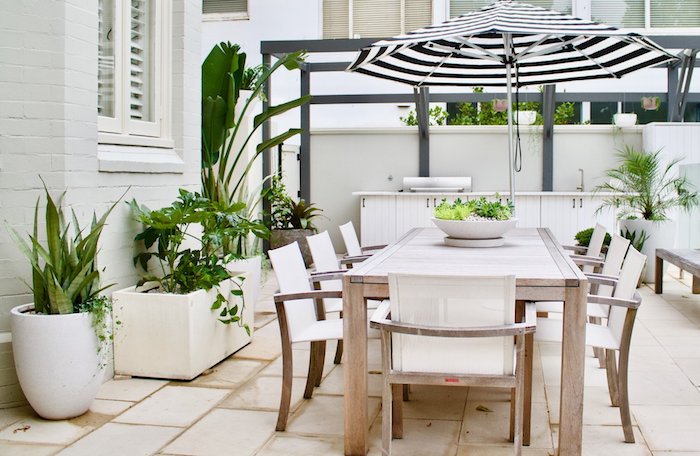 salle à manger extérieure en table et chaises bois, carrelage beige, plantes en pots et bacs, pergola moderne grise, parasol noir et blanc