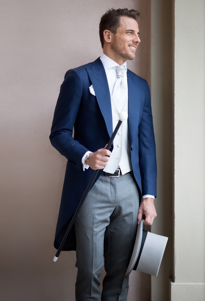 un modèle de redingote homme dépareillée avec pantalon gris, une veste bleu marine profond, gilet blanc et lavalière vintage
