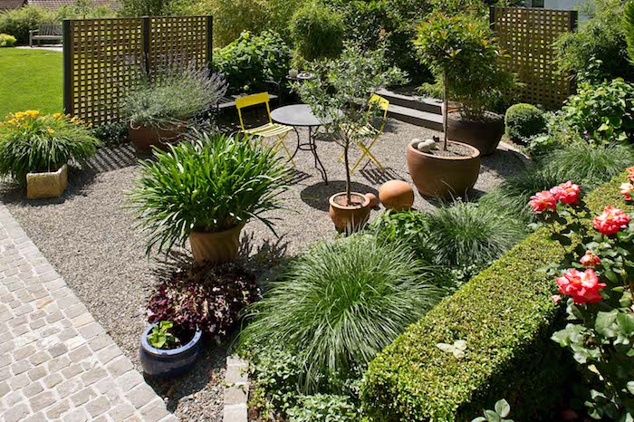 chemin de pavage exterieur, terrasse recouverte de gravier, plantes en pots, bordure de buis, chaise et tables metalliques, treillis occulants