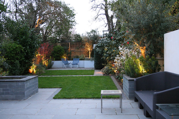 dalles de béton et carrés de gazon sur une terrasse exterieure, chaises metalliques, bordure de végétaux, canapé noir