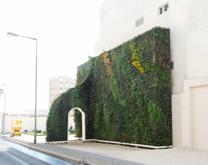mur vegetal exterieur avec un arc avec une porte blanche, installation en vert, jaune et marron, culture verticale, élément complémentaire d'un immeuble aux murs blancs 