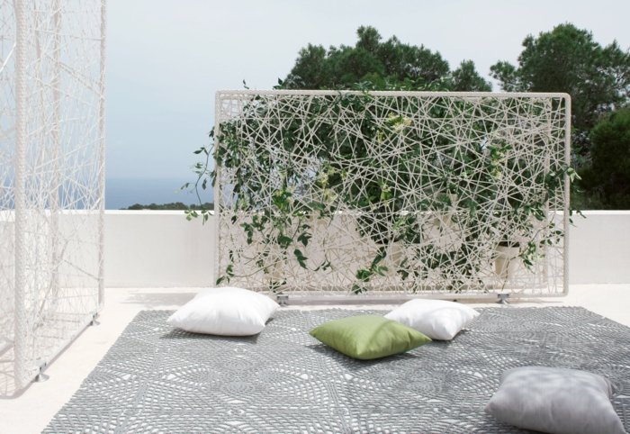 mur vegetal exterieur sur un panneau blanc aux fils blancs qui supportent le lierre, installation sur une terrasse avec un grand tapis gris et des coussins en vert, blanc et gris