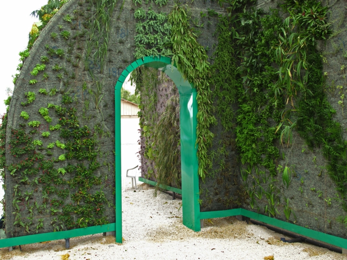 jardin vertical, arc avec la partie intérieure en bois peint en vert herbe, 4 types de plantes vertes qui ornent les murs, parc public, sol recouvert de sable 