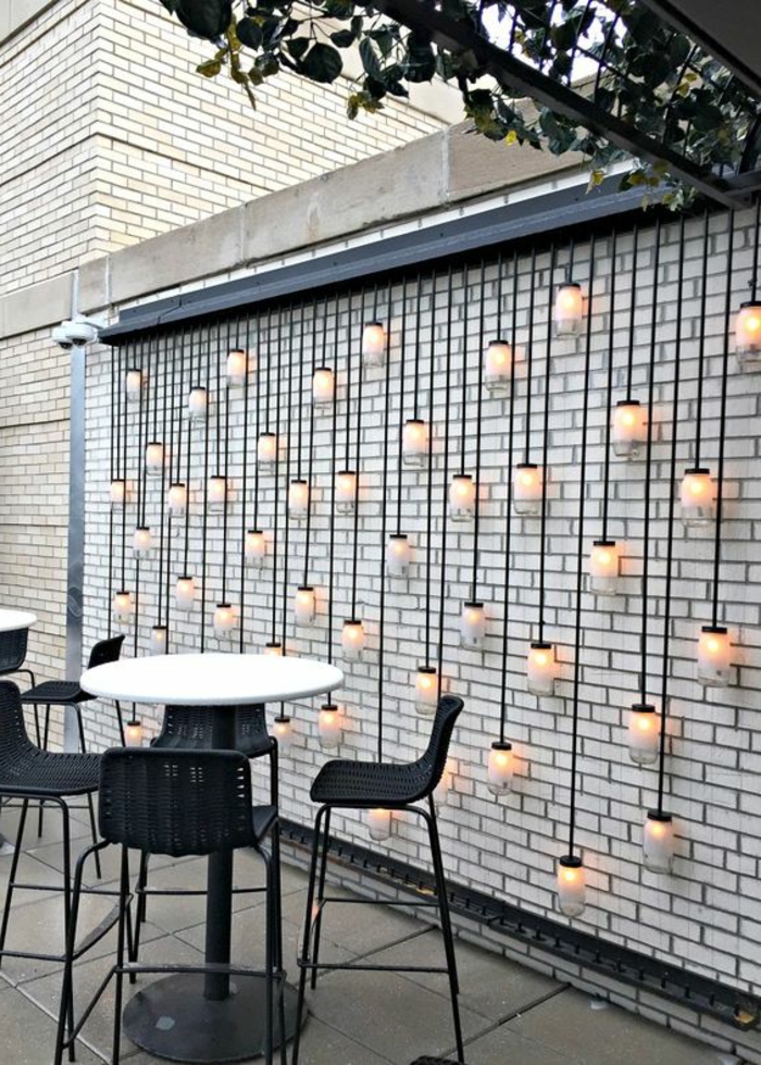 idee deco jardin exterieur avec des grandes ampoules suspendues tout au long d'un mur en briques blanches, table haute et ronde avec plan blanc et pieds en métal noir, trois chaises entièrement en noir, en PVC et métal