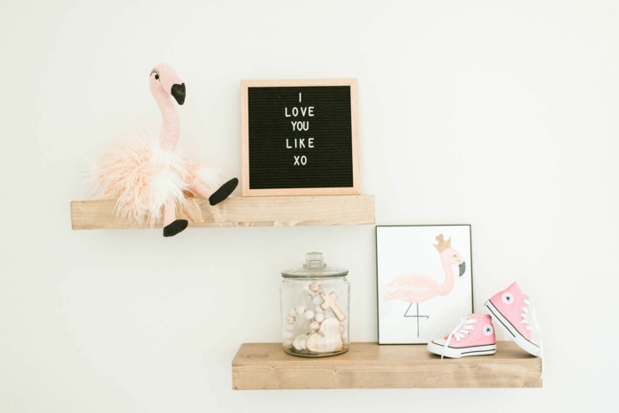 accessoire flamant rose, étagères en bois clair sur un mur blanc crème dans une chambre de bébé, tableau décoratif avec message d'amour, petits baskets roses, bocal en verre avec un cœur et une croix décoratives