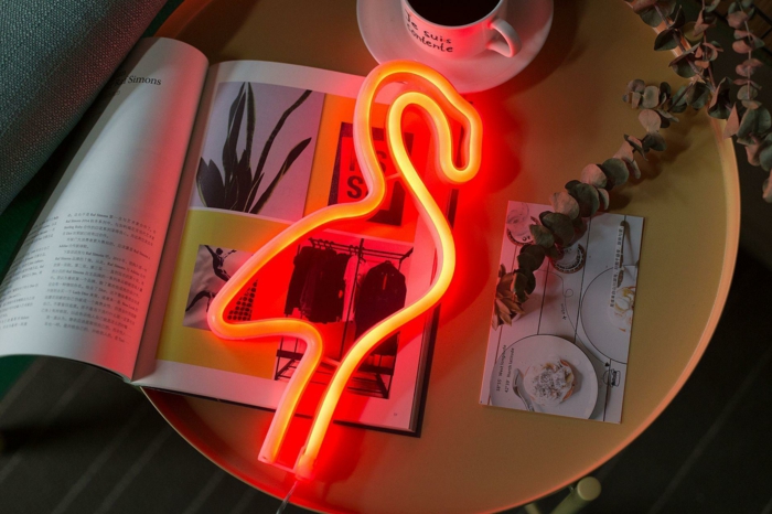 lampe néon en forme de flamant lumineux, petite table ronde de chevet avec un magazine de mode, carte postale et petite tasse de café blanche, objet deco original, déco flamant rose