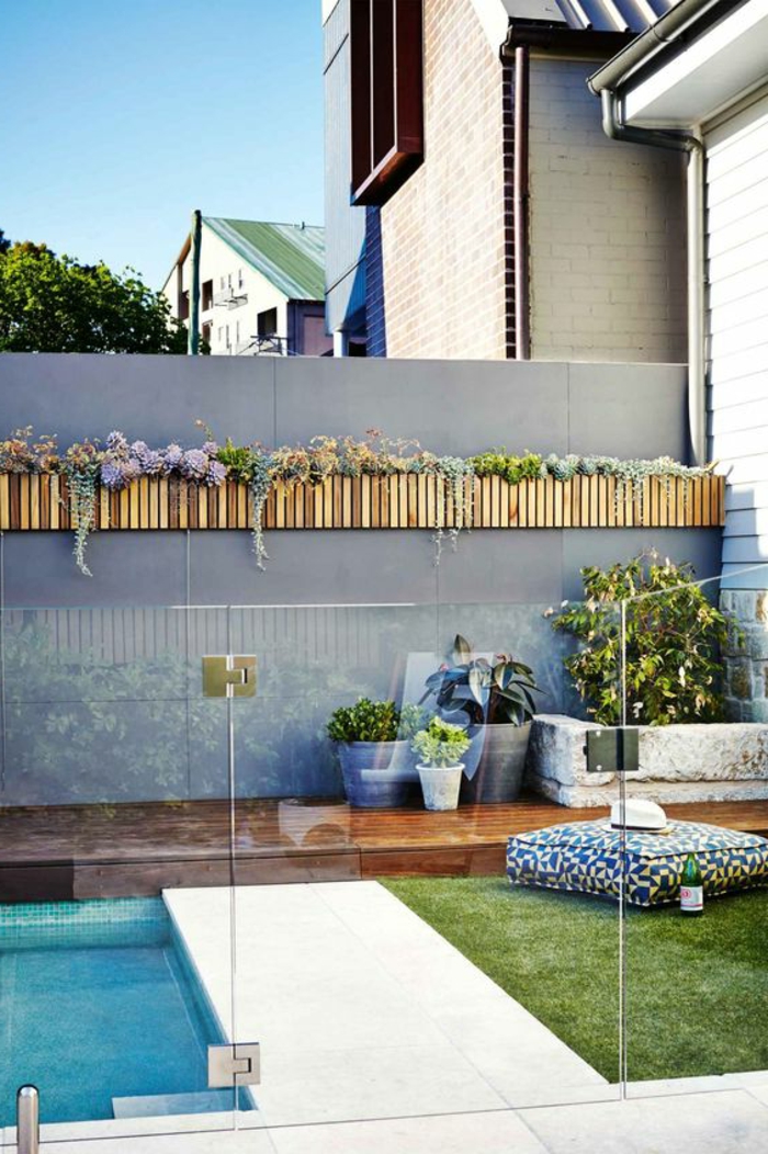 deco mur exterieur, habillage mur exterieur avec des porte-plantes en bois clair, clôture de jardin avec piscine en couleur gris pastel, herbe verte avec un gros coussin carré pour relaxer