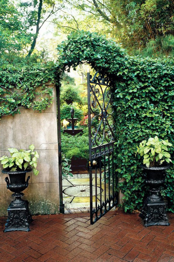 revetement mural exterieur, jardin classique en style vintage, habiller un mur extérieur avec des plantes rampantes vertes, grande porte en métal noir, deux vases noirs classiques avec des poignets des deux cotes