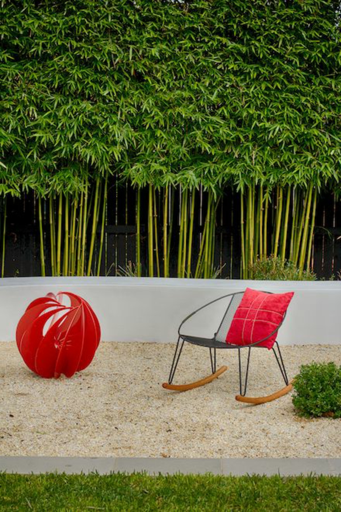 cloison végétale avec des tiges de bambou, coin de jardin relax avec un fauteuil en métal noir et bois clair, boule en tissu rouge, décoration design, sable fin, herbe verte bien entretenue, mur vegetal exterieur 