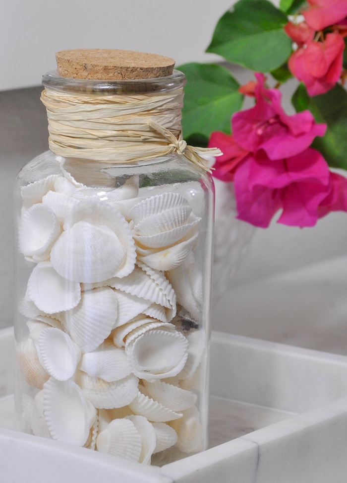idée d activité manuelle ado avec fiole en verre remplie de petites coquilles blanches dans une assiette blanche, fleur fuchsia à coté
