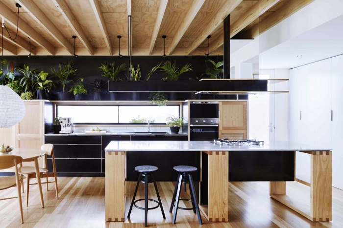 aménagement de cuisine moderne en bois avec plafond en poutres de bois clair et pan de mur en noir mate, décoration avec plantes vertes