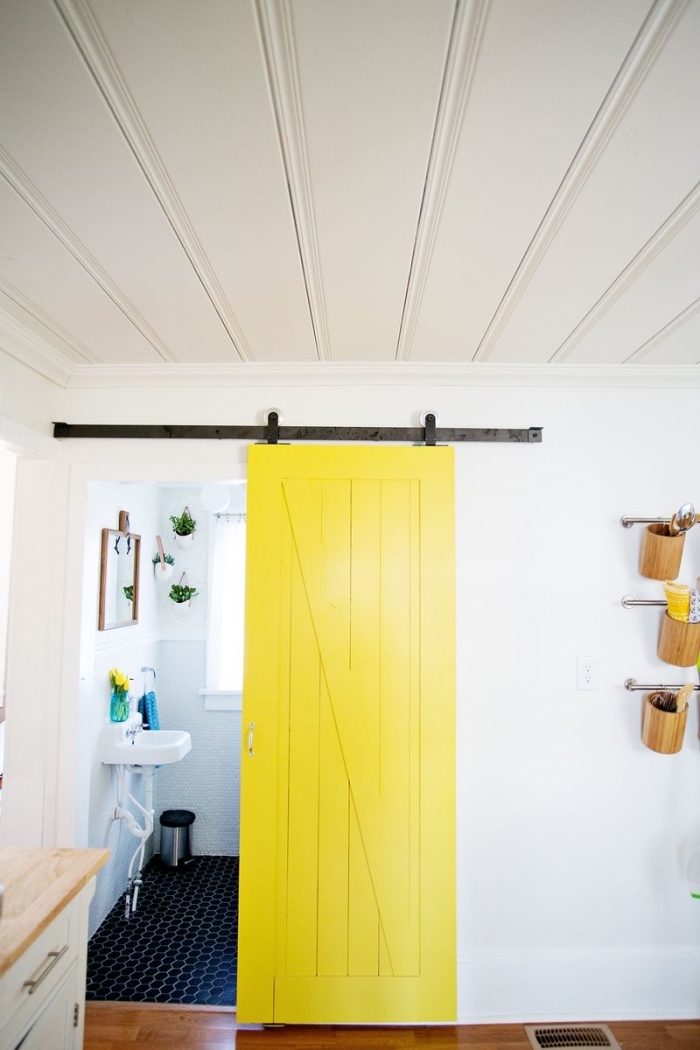 une porte interieur bois de grange repeinte en jaune fluo qui délimite la salle de bain et contraste avec les mures blancs et le carrelage noir