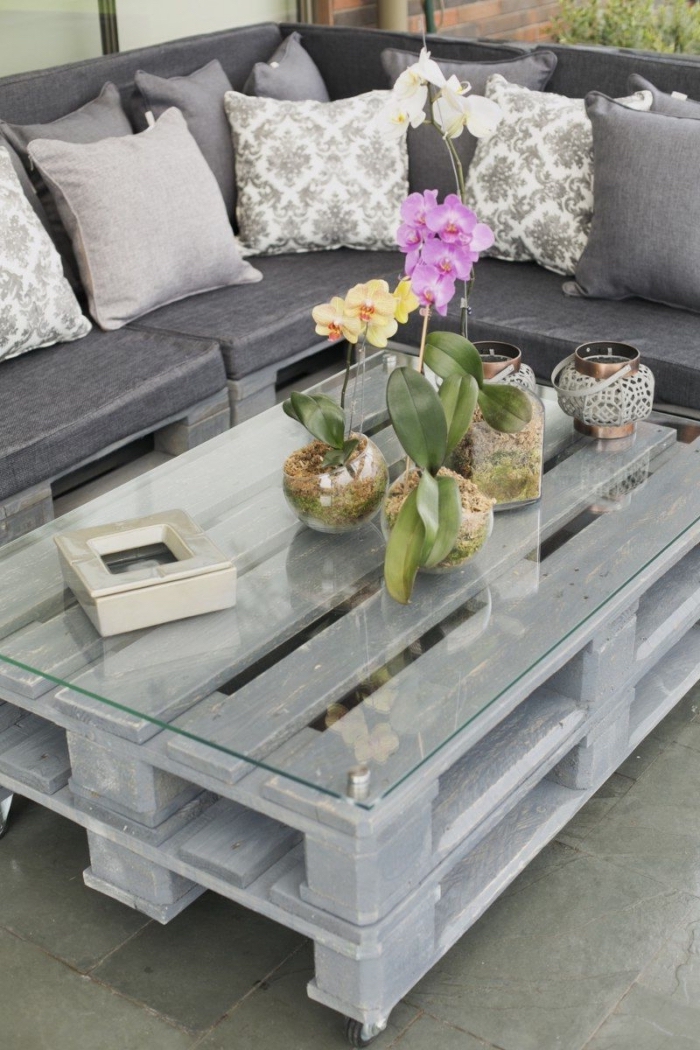 une table basse exterieur patinée grise avec un plateau en verre et une jolie déco d'orchidées en vase en verre