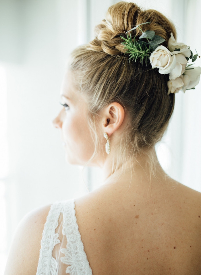 chignon-tressé-mariage-avec-un-chignon-haut-entouré-de-tresse-et-des-gleurs-blanches-robe-de-mariée-blanche chignon tressé haut, exemple de chignon classique entouré de tresse et une attache en fleurs fraiches