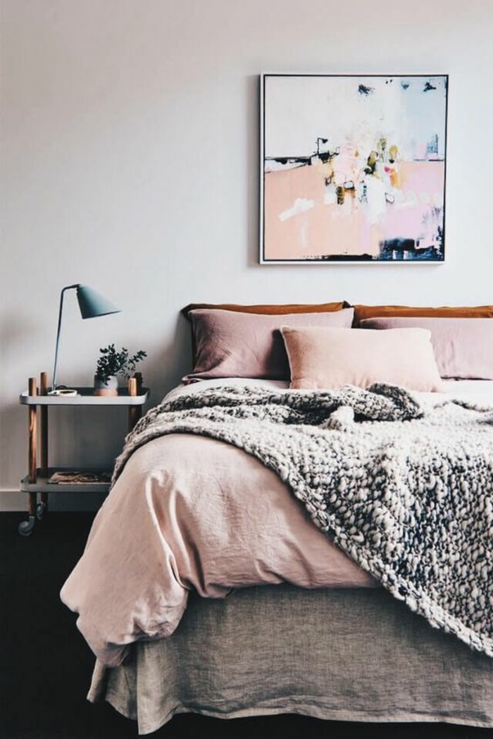 couverture vieux rose dans une chambre d'adulte, tableau art abstrait au-dessus du lit, couverture en grosse maille grise, coussins en couleur rose pale 