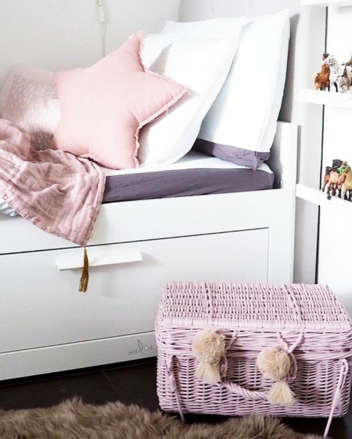 chambre avec lit en bois peint en blanc, coussin en couleur rose pale en forme d'étoile, deux coussins blancs, malle en rotin tressé peinte en rose avec deux pompons décoratifs devant