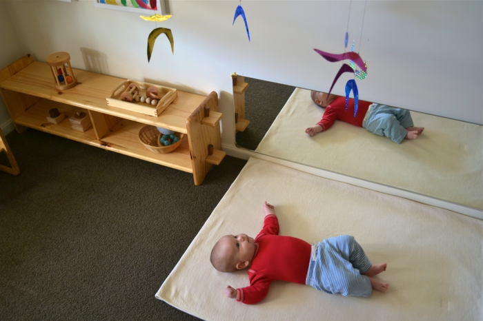 miroir montessori dans la chambre de bébé, meuble montessori, meuble de rangement en bois clair, moquette en marron foncé, bébé qui contemple un jouet mobile suspendu au plafond sur un fil transparent 