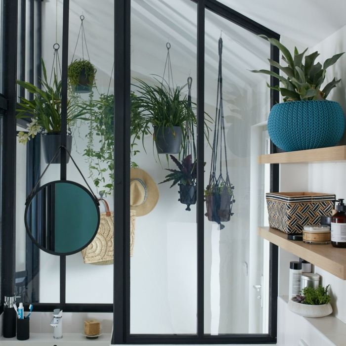 cloison verriere en métal noir avec du verre transparent, étagères en bois clair, porte-plantes suspendues en macramé, miroir rond suspendu avec lavabo blanc 