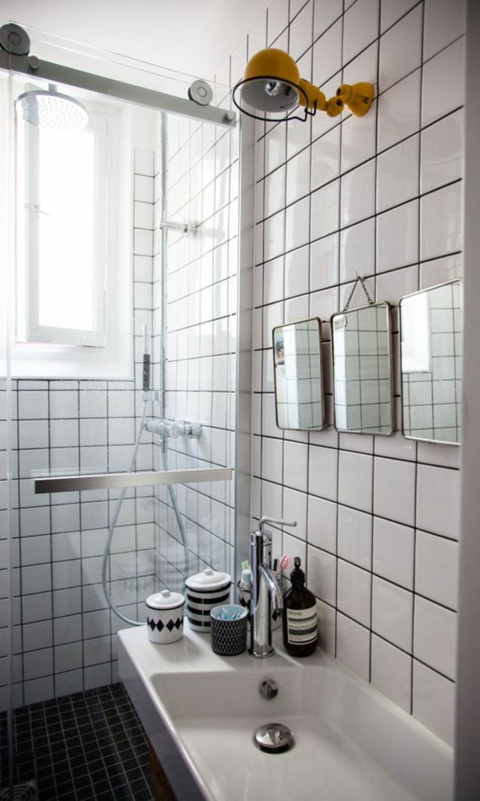 salle de bain avec verrière, luminaire en métal jaune sur le mur au-dessus du lavabo blanc carré, carrelage blanc, verriere douche en métal couleur argent, trois miroirs carrés au-dessus du lavabo 