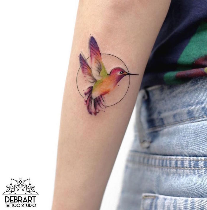 petit tatouage de colibri en couleurs aquarelle dans cercle noir sur avant bras de femme par debrart