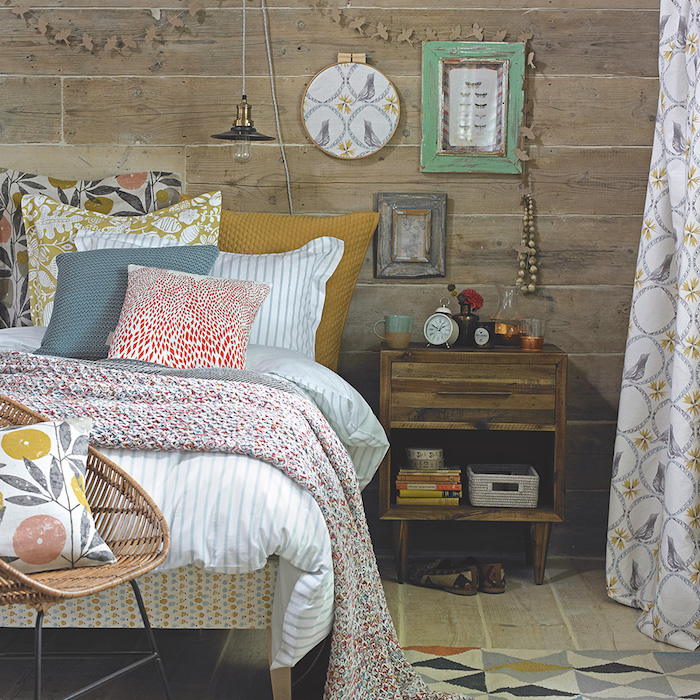 murs et sol en bois, linge de lit et coussins à motifs fleuris, table de nuit en bois brut décorée d objets vintage, chaise en rotin