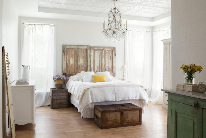 campagne decoration vintage avec tete de lit en portes bois granges, coffre vintage en guise de bout de lit, table de nuit bois brut, linge de lit blanc, parquet clair, lustre baroque
