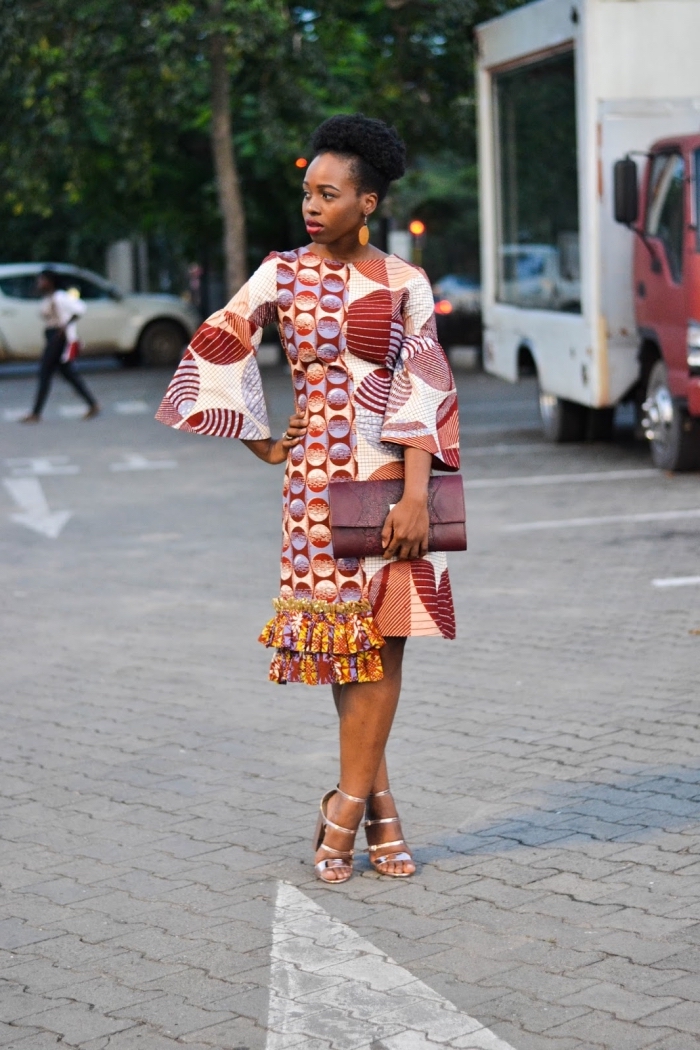 modèle de robe africaine moderne et chic avec manches trois-quart évasées, à motifs tribaux dans les tons ocre accessoirisée avec une pochette en cuir bordeaux