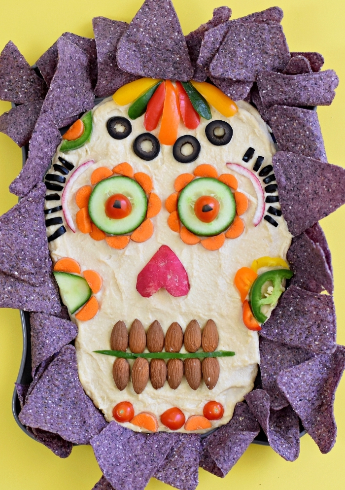 recette de houmous d'halloween fait-maison décorés avec des morceaux de légumes, d'amandes et d'olives pour former une tête de mort mexicaine