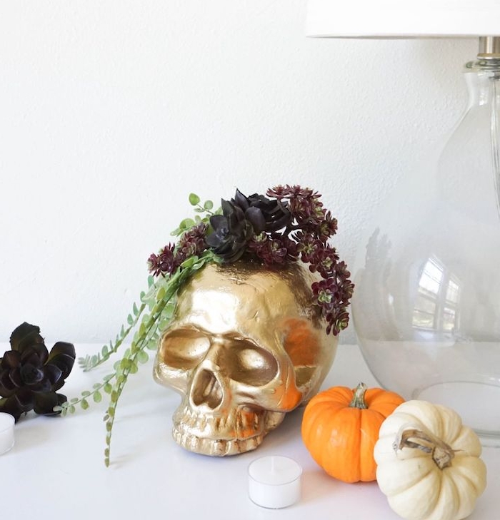 crâne décorative couleur or avec decoration de plantes, entourée de potirons décoratifs, bougie et fleur artificielle noire