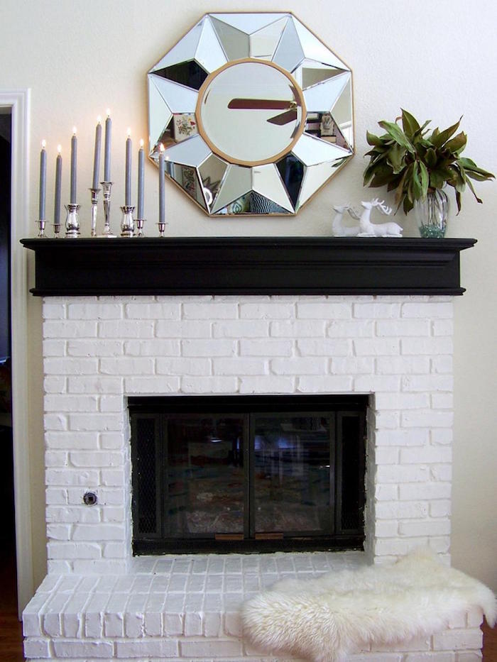 cheminée avec manteau en briques repeintes en blanc et support noir mat avec chandelier et bougies, miroir chrome design sur mur blanc