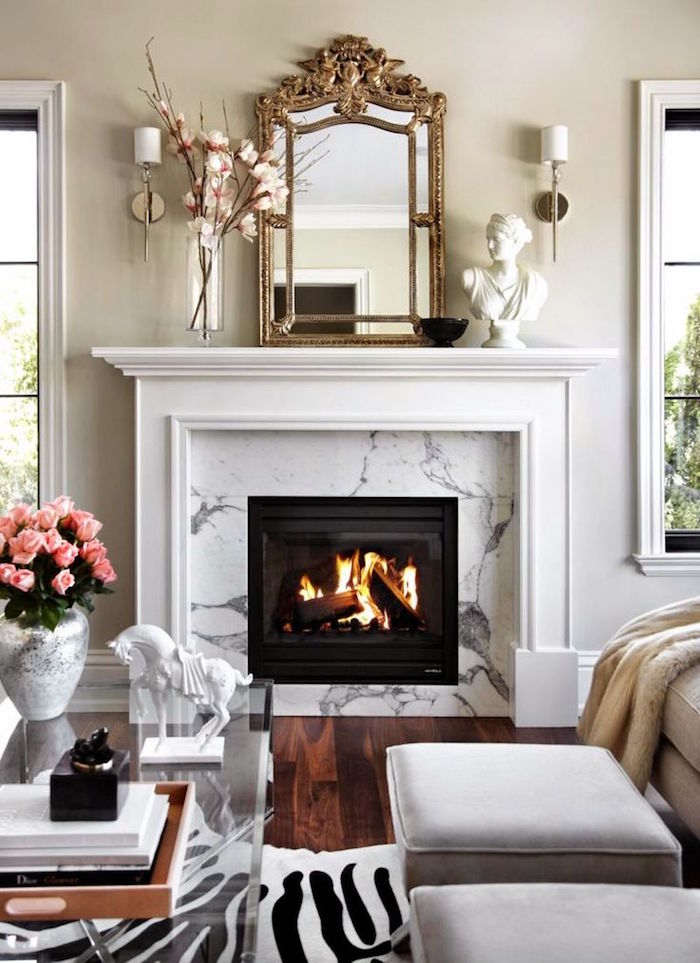 manteau de cheminee blanc classique avec cadre marbre blanc gris, salon avec mur blanc cassé, sol parquet foncé, objets déco avec statuette et miroir doré vintage