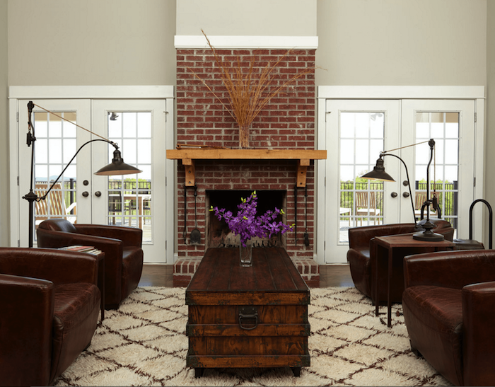 salon à déco vintage avec fauteuils en cuir foncé, table en bois ciré type malle et manteau de cheminée en briques rouges avec étagère en bois