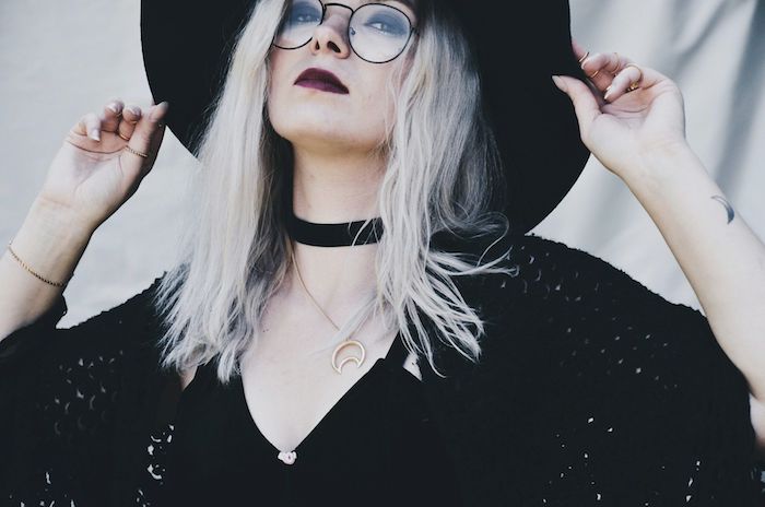accessoire de sorcière collier ras du cou, gilet à grosses mailles noire, lunettes de vue monture ronde, coloration blond platine, rouge à lèvres rouge, maquillage sorciere halloween