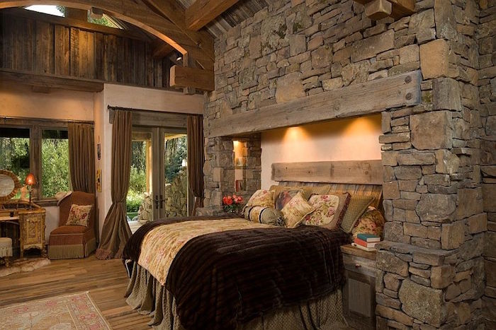 chambre style chateau moyen age avec murs en vieilles pierres grises différentes formes et lit dans cadre de cheminée et charpente en bois