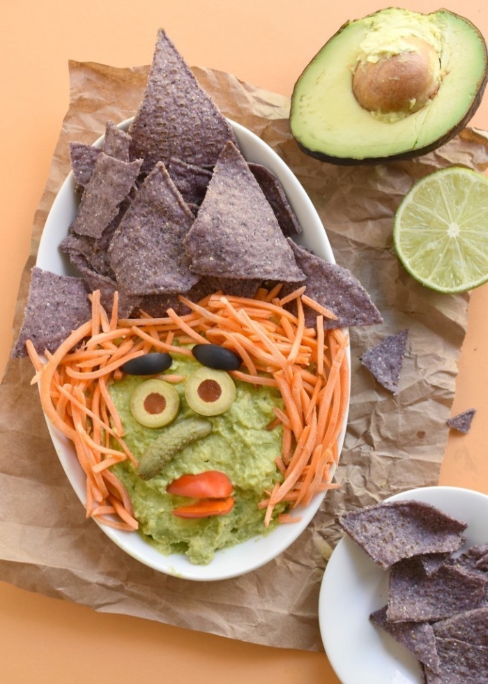 guacamole pour l'apéritif d'halloween en forme de tête de sorcière décoré avec chips tortilla de sarrasin, olives, carottes râpées et mini-cornichon