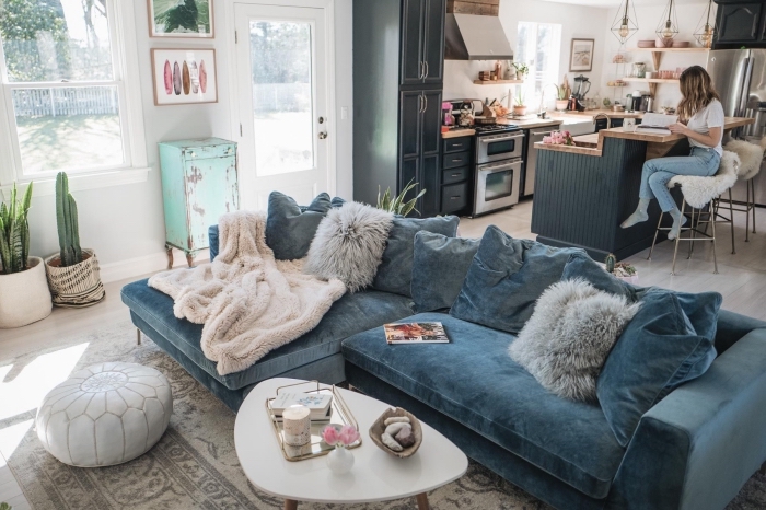 un grand canapé cosy en velours couleur bleu canard qui trône au centre d'un salon bohème chic ouvert sur la cuisine