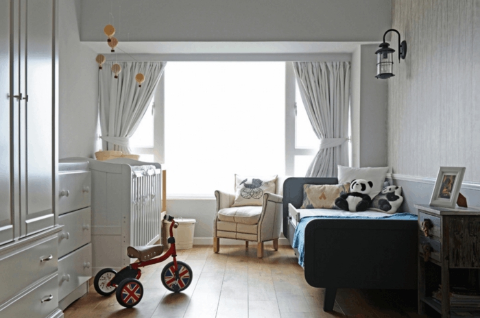 rideaux en gris clair, vélo à trois roues, lit enfant 2 ans, sol en planches, fauteuil crème, lanterne murale