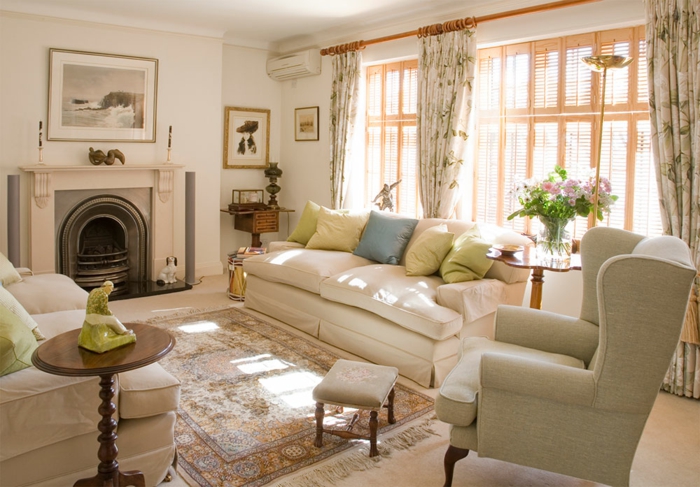 salon clair et lumineux, sofas blancs, tapis persan, grand fauteuil gris, cheminée murale blanche, rideaux floraux