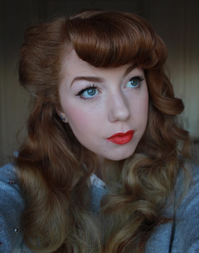 femme rousse teint pale avec maquillage années 50 lipstick et rose à joues et coiffure de pin up avec frange enroulée et boucles victory rolls