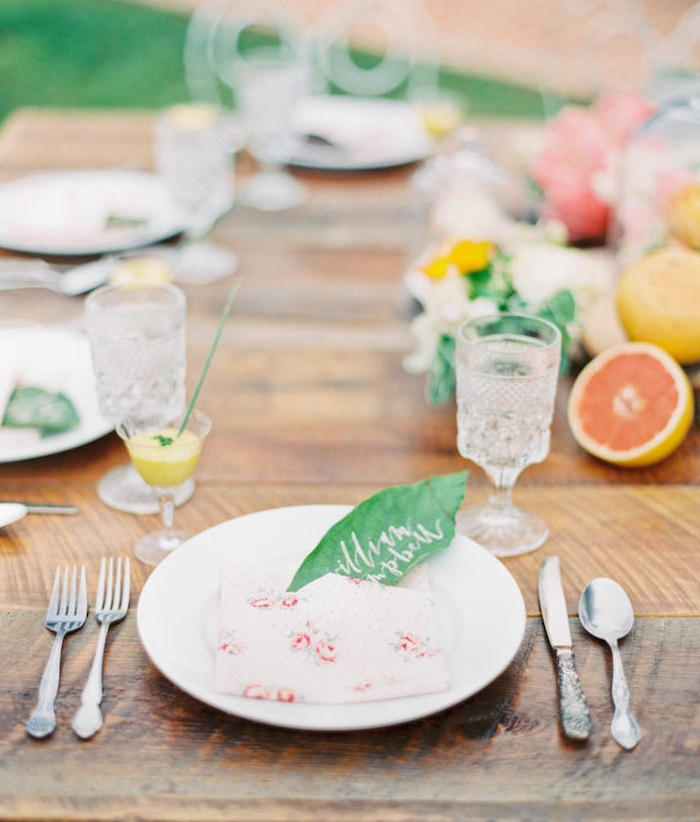 idee de table style champetre chic mariage avec centre de table en fleurs et fruits frais, serviette pliée avec poche et marque place en feuille verte avec nom invité sur table bois brut