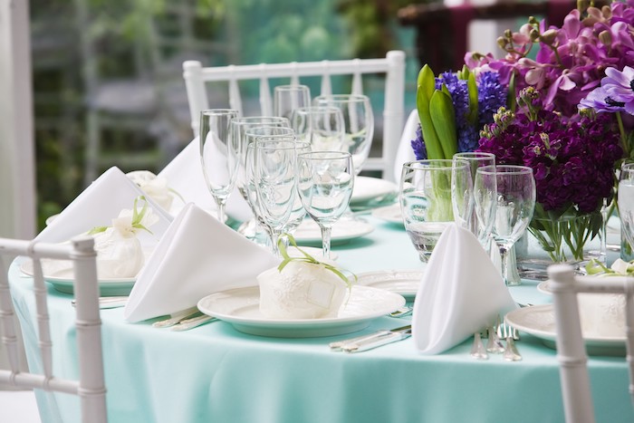 décoration élégante de mariage avec des serviettes blanches pliés en triangles sur nappe bleu clair, verres à vin, composition florale en fleurs mauve et violettes