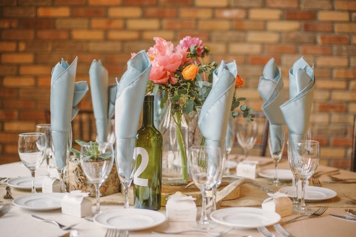 serviettes en tissu grises dans des verres à vin, nappe blanches, assiettes blanches et centre de table bouquet de fleurs dans une bouteille en verre recyclée, numéro de table écrit sur bouteille