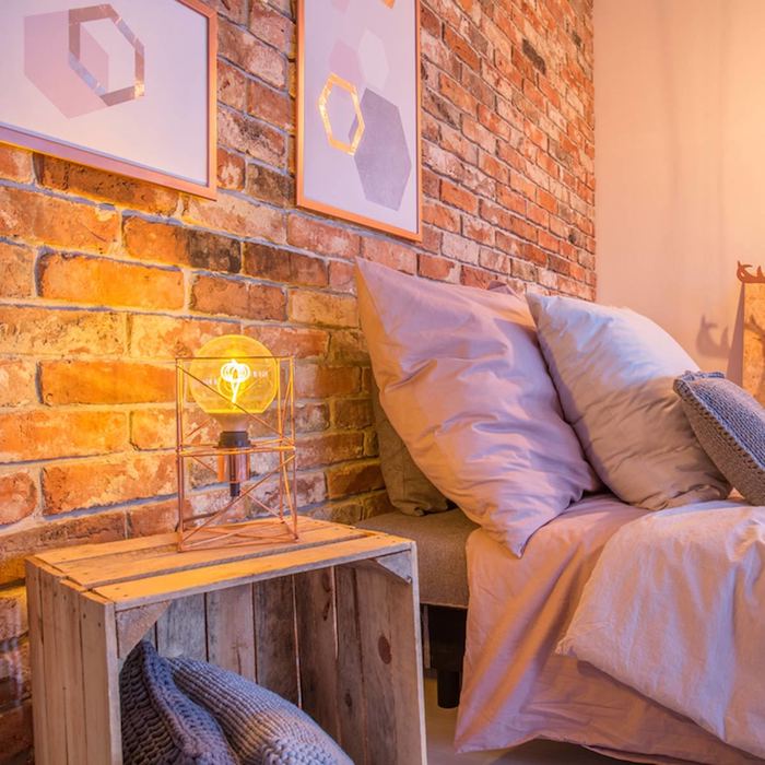 table en cagette bois brut avec des coussins rangés à l intérieur et une lampe ampoule dessus, mur de briques, linge de lit gris