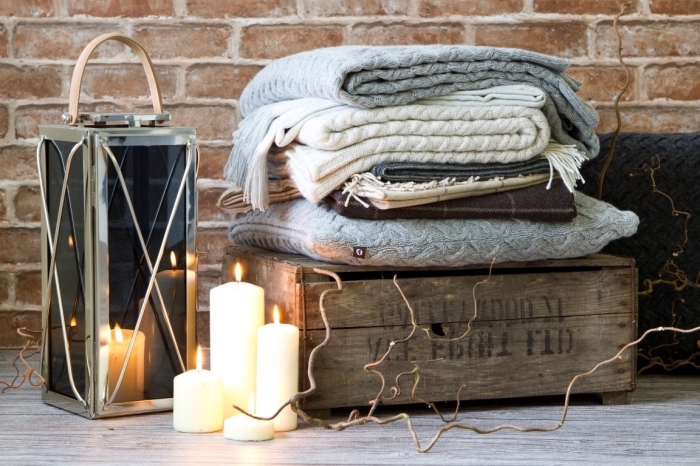 image cocooning avec une pile de plaids douillets sur une caisse en bois vintage, une lanterne déco et quelques bougies allumées