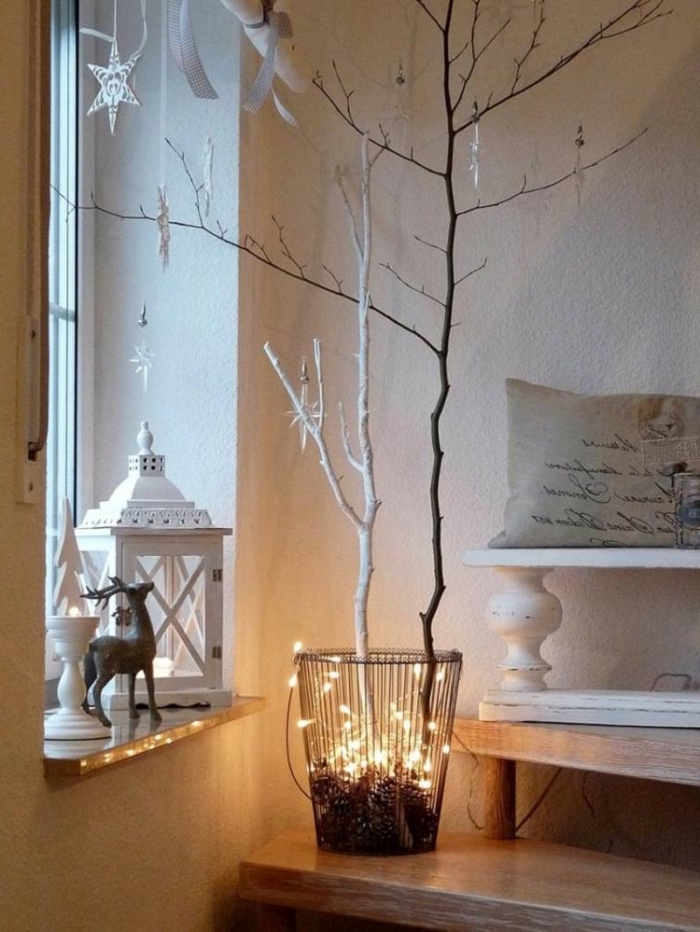 décoration rebord de fenêtre avec une lanterne en métal blanc, un bougeoir et une figurine de cerf, décoration de noel à fabriquer en bois, branches d'arbre en corbeille décorées avec une guirlande lumineuse