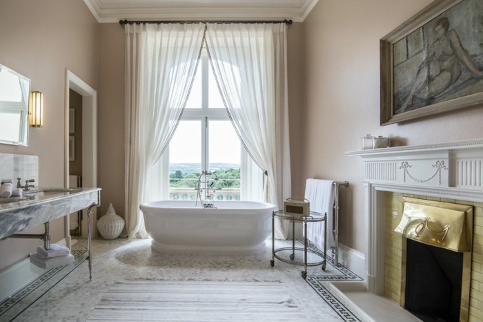salle de bain spacieuse, tapis rayé couleurs claires, rideau blanc, grand lavabo marbré, cheminée, portrait de femme encadré