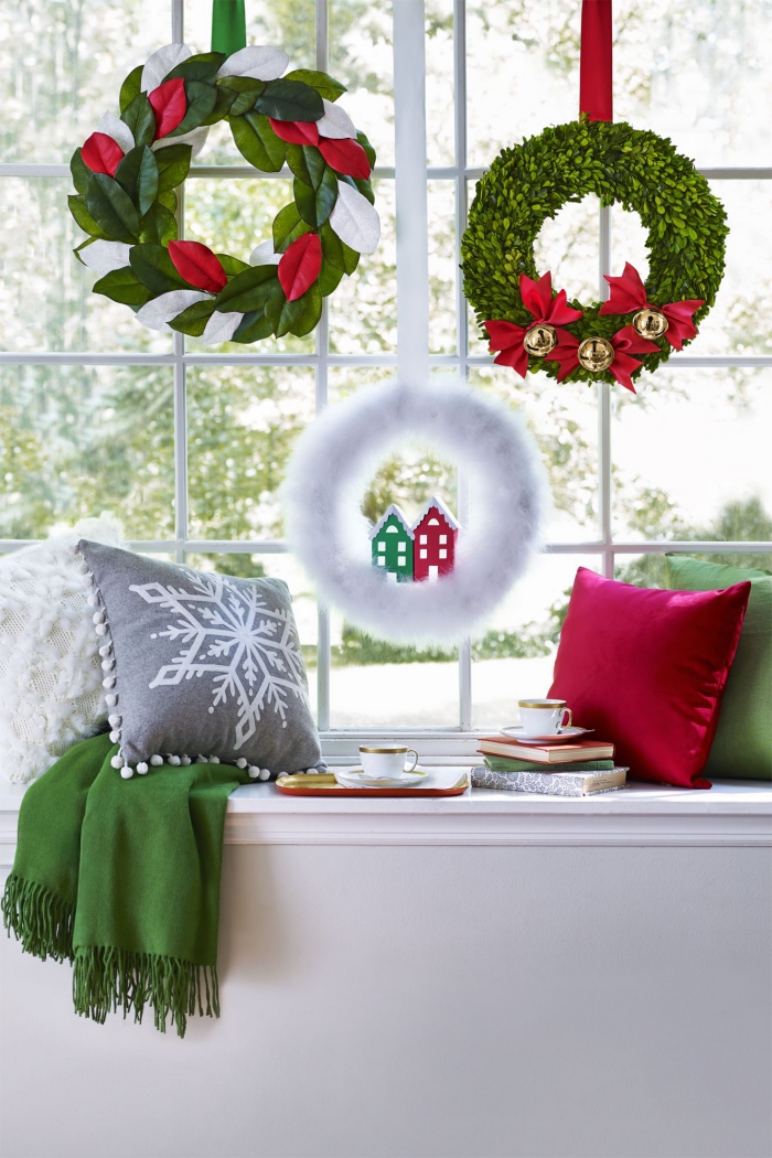 trois couronnes de noël aux couleurs traditionnelles de la fêtes suspendues à la fenêtre avec un rebord décorés avec coussins blanc, rouge, vert et gris transformé en coin lecture cosy