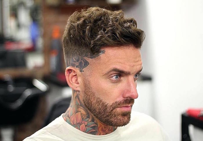 hipster homme avec tatouage fleur lotus dans le cou et coupe dégradé court avec tatouage sur la tempe et dessus plus long avec cheveux ondulés et yeux bleus